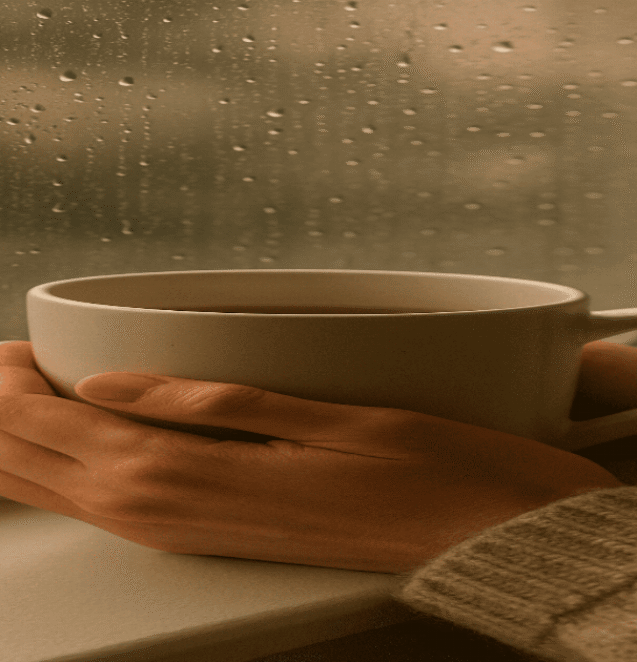 Cozy close-up of hands holding a warm mug near a window with raindrops, with soft lighting and a peaceful atmosphere