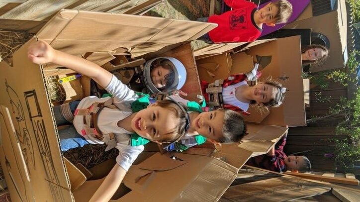 A toddler joyfully exploring inside a DIY cardboard spaceship alongside other children