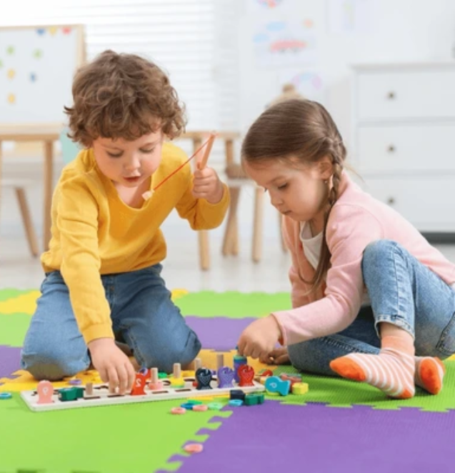 Two young children playing on a colorful foam mat inside a cozy, safe indoor space filled with soft blocks and engaging toys