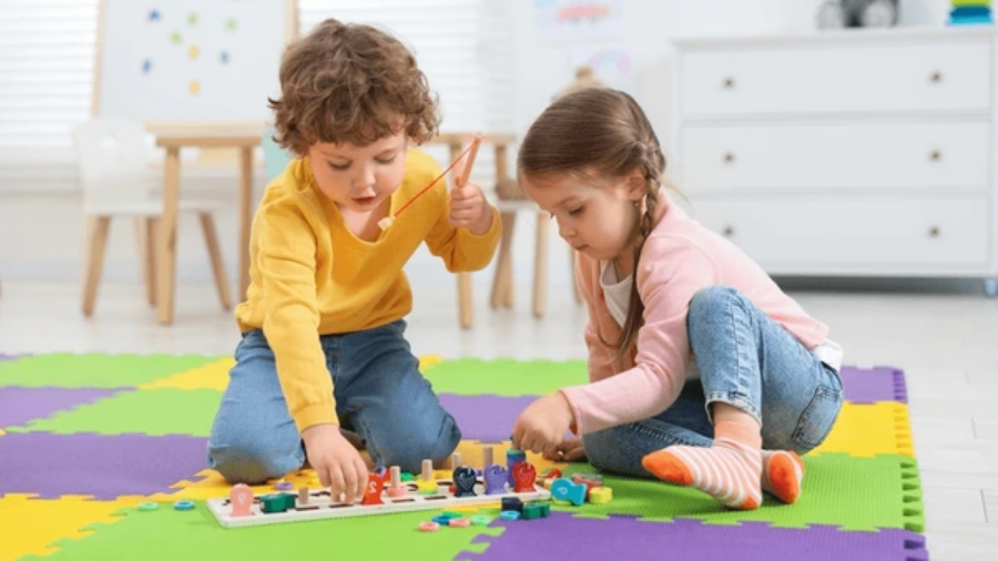 Two young children playing on a colorful foam mat inside a cozy, safe indoor space filled with soft blocks and engaging toys